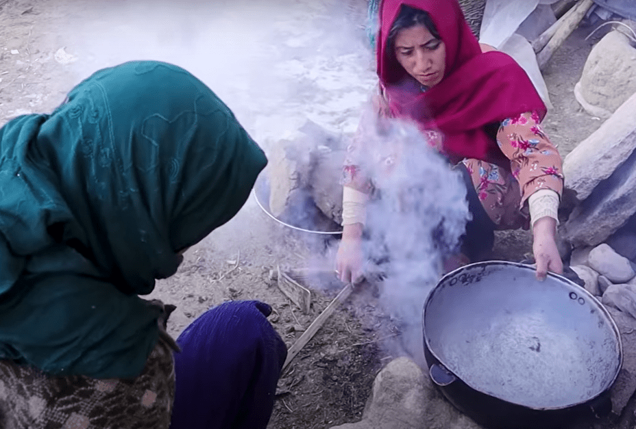 Dues dones cuinant amb una olla sobre una font de foc, envoltades de fum, en un entorn rural.